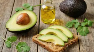 Fette di avocado su pane integrale con mezzo avocado, olio e foglie verdi su tavolo di legno